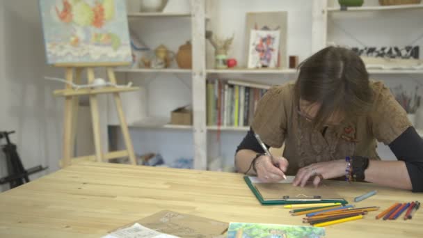 Женщина рисует картинку или открытку сидя за столом с красочными карандашами — стоковое видео