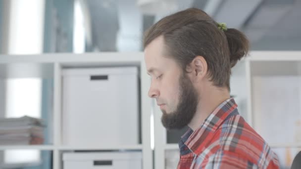 Un uomo con la barba e un ciuffo di capelli in testa, lavora in ufficio . — Video Stock