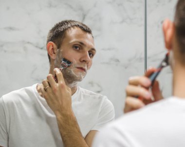 Genç bir adam aynanın yanında sakalını kesiyor.