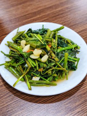 Beyaz bir tabakta servis edilen sarımsak ve dilimlenmiş kırmızı biberle pişirilmiş karıştırılmış su ıspanağı. Sağlıklı sebze menüsü. Vejetaryen mutfağı. Yemek menüsü görüntüsü.