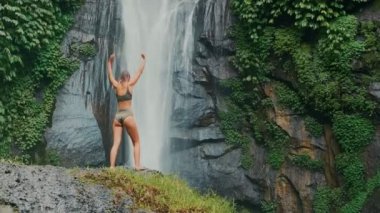 Şelalenin önünde ellerini kaldırmış genç bir kadının dikiz görüntüsü..