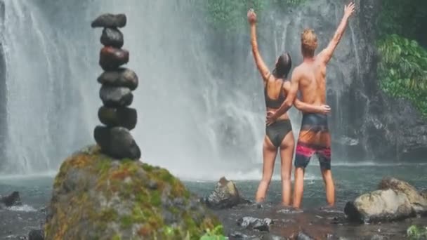 Jovens viajantes felizes ficam em frente à cachoeira com as mãos levantadas — Vídeo de Stock