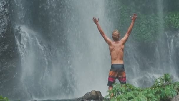 Homem na cachoeira levantando as mãos em se sentir mais perto da natureza. Viajar para Bali. — Vídeo de Stock