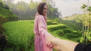 Genç bir kadın el ele tutuşup pirinç tarlalarının ortasında pembe kimono giyerek yürüyor.
