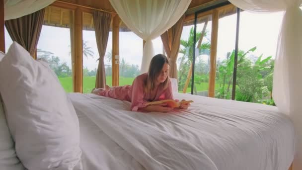 Молодая женщина читает книгу, лежащую в постели прекрасного бамбукового дома посреди поля — стоковое видео