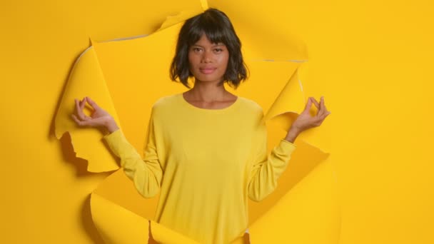 Relaxed young dark skinned woman makes mudra gesture through torn yellow wall — Stock Video