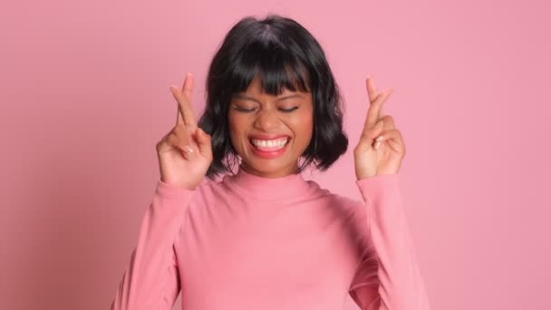 Бодрая темнокожая женщина скрещивает пальцы на удачу на розовом фоне — стоковое видео