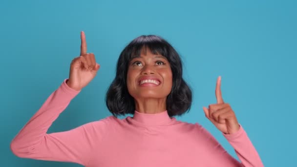 Positive woman in casual clothing points index fingers up over blue background — Stock Video