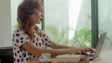 Kulaklıklı kadın çağrı merkezi çalışanı dizüstü bilgisayara bakar ve not defterine yazar.