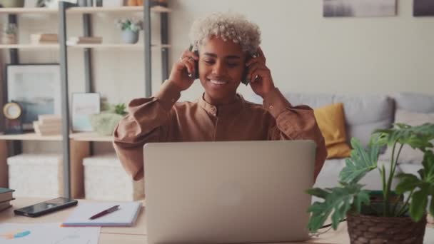 Mujer africana sonriente poniéndose auriculares inalámbricos, comenzando el día de trabajo remotamente — Vídeos de Stock