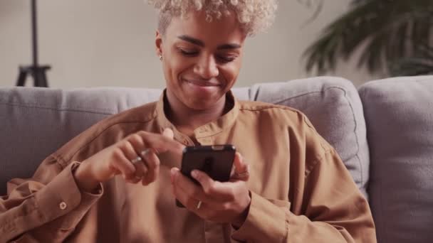 Улыбающаяся африканская женщина отдыхает на уютном диване, используя смартфон, полученное сообщение — стоковое видео