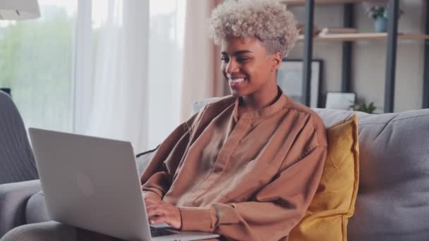 Jovem mulher africana feliz sentado no sofá usando laptop olhando para a tela — Vídeo de Stock