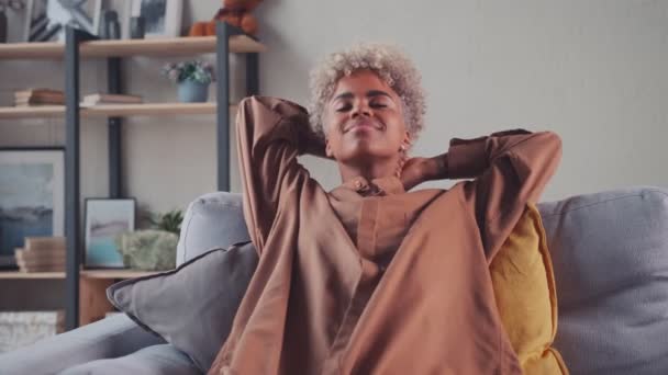 Африканская женщина отдыхает на диване, делая глубокий вдох, держа руки за голову — стоковое видео