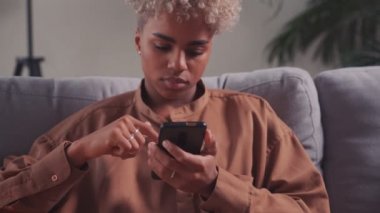 Akıllı telefonuyla kanepede dinlenen Afrikalı kadın modern kablosuz cihazın tadını çıkarıyor