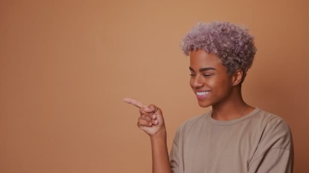 Dark skinned woman with appealing smile pointing on left side over brown wall. — Stock Video