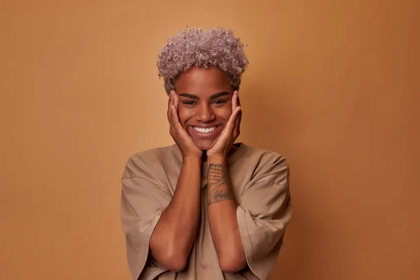 Mujer africana sorprendida con el pelo rubio toca las mejillas de ambas manos con sonrisa. — Foto de Stock