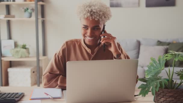 Ev ofisinde dizüstü bilgisayarla oturan Afrikalı bir kadın cep telefonuyla konuşuyor. — Stok video