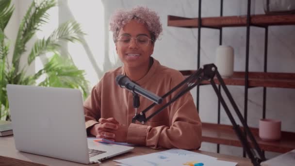 Веселая африканская женщина записывает подкаст из дома с ноутбуком и микрофоном — стоковое видео