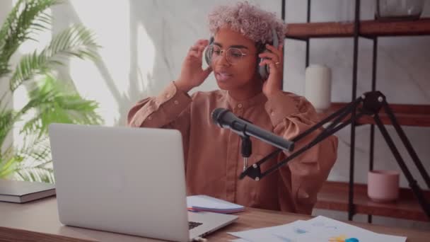 Czarna kobieta z blond kręconymi włosami zakłada słuchawki i rozpoczyna audycję radiową. — Wideo stockowe