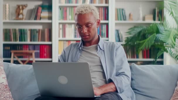 Boldog sötét bőrű férfi ül a kanapén laptoppal sikoltozva igen felemelt ököl — Stock videók