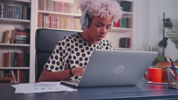 Vrouw luistert naar muziek in koptelefoon aan het bureau met behulp van laptop dansen in goed humeur. — Stockvideo