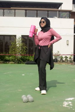 Genç Asyalı Müslüman kadın tesettür takıyor ve parkta mineral pembe şişe tutuyor.