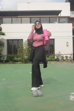 Genç Asyalı Müslüman kadın tesettür takıyor ve parkta mineral pembe şişe tutuyor.