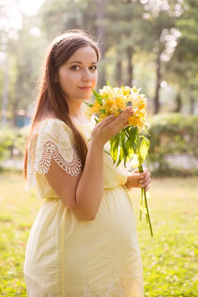 A bela mulher grávida jovem com flores no parque no dia de verão — Fotografia de Stock