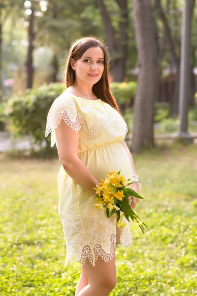 Die junge schöne schwangere Frau mit Blumen im Park an einem Sommertag — Stockfoto