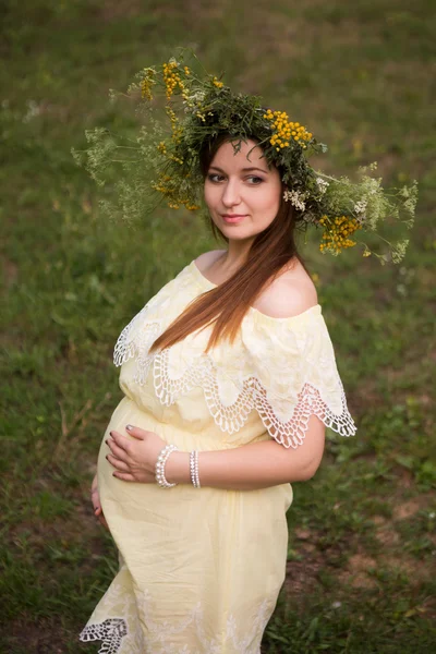 De mooie zwangere vrouw in een krans van bloemen buiten in de natuur — Stockfoto