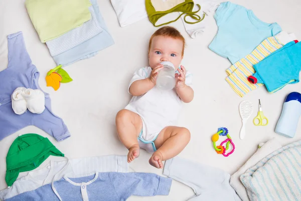 The baby drinks from a small bottle. Baby on white background with clothing, toiletries, toys and health care accessories. Wish list or shopping overview for pregnancy and baby shower. — Stock Photo, Image