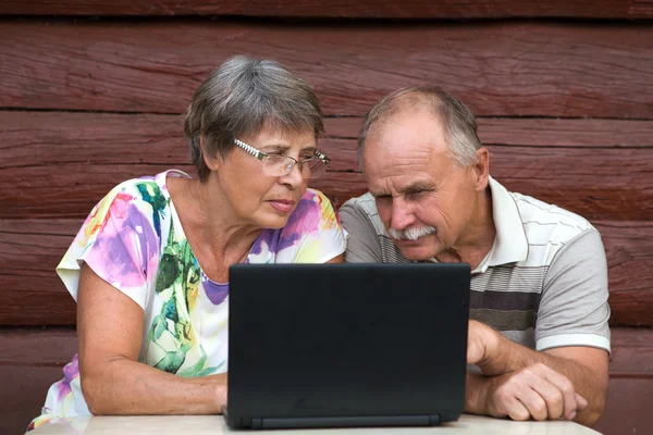 Пожилая пара смотрит на свой ноутбук на деревянном фоне дома — стоковое фото