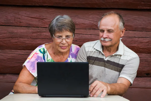 Пожилая пара смотрит на свой ноутбук на деревянном фоне дома — стоковое фото