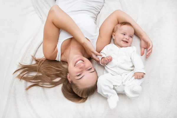 Mãe e filho em uma cama branca. Mãe e bebê em fralda brincando no quarto ensolarado . — Fotografia de Stock