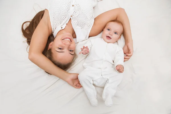 母亲和孩子在一张白色的床上。妈妈和婴儿的尿布在阳光明媚的卧室里玩. — 图库照片