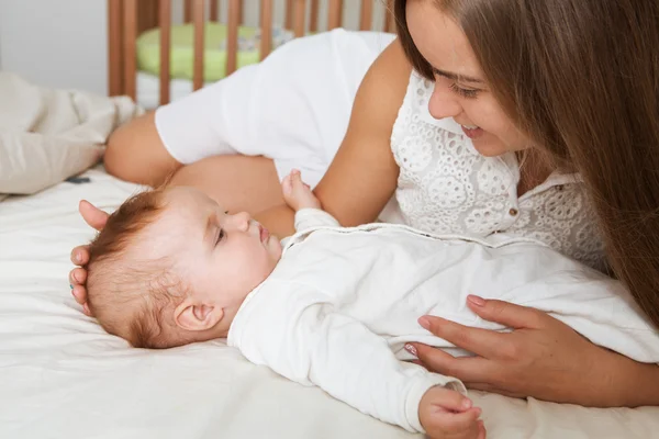 Anne ve çocuk beyaz bir yatakta. Anne ve bebek bezi güneşli yatak odasında oynarken.