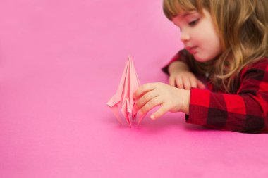 Küçük kız pembe arka planda origami kağıt uçağıyla oynuyor.