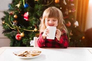 Noel ağacının yanındaki masada oturmuş sıcak çikolata içen tatlı küçük kız.