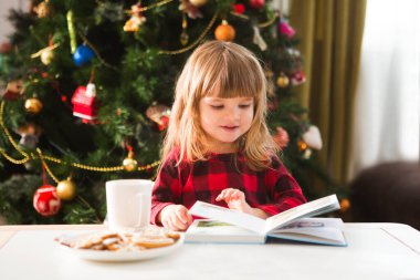 Küçük tatlı kız kitap okuyor, Noel ağacının yanındaki masada oturuyor.