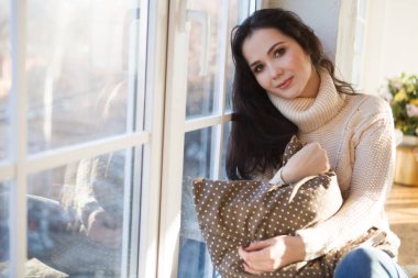 Romantik esmer kadın, süveter ve kot pantolon içinde, pencerenin kenarında oturuyor.