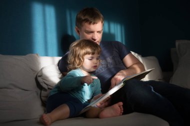 Baba ve kızı kitap okurken dairede kanepede oturuyor. 