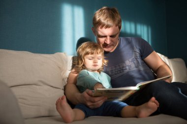 Baba ve kızı kitap okurken dairede kanepede oturuyor. 