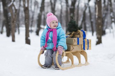Güzel, küçük bir kız, kızağın yanında duruyor. Süslü yeni yıl ağacı ve karlı ormanda Noel kutuları var.