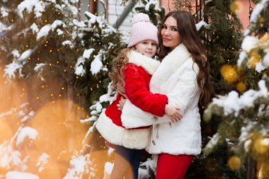 Güzel küçük bir kız ve annesi kalın giysiler içinde karla kaplı köknar gibi poz veriyorlar. 