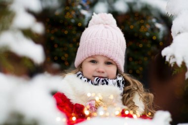 Karlı ormanda, sıcacık giysiler içinde, köknar ağaçlarının yanında duran tatlı küçük bir kız.