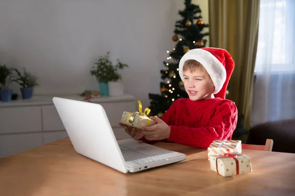 Happy child  in a Christmas hat with a Christmas gift using a laptop communicates with their friends on video call using webcam.  stays at home for Christmas holidays.