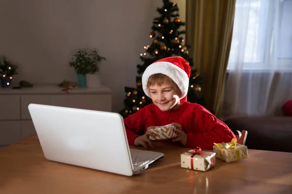 Happy child  in a Christmas hat with a Christmas gift using a laptop communicates with their friends on video call using webcam.  stays at home for Christmas holidays.