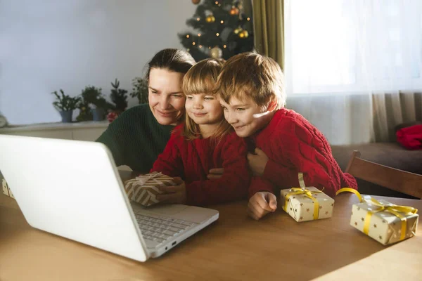Family mom and two kids with a Christmas gift using a laptop communicates with their friends on video call using webcam.  stays at home for Christmas holidays.