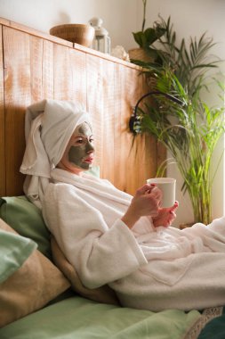 Kafasında bornoz ve havlu olan bir kadın evinde dinlenen bir fincan çayla yüz maskesi takıyor. organik cilt bakımı. Yaşam tarzı. Güzellik kaplıcası ve kozmetoloji. 