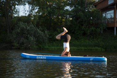 Yemek masasında yoga yapan bir kadın gün batımında kürek tahtasında amuda kalkıyor. Açık hava yaz aktivitesi. N 'aber yoga.